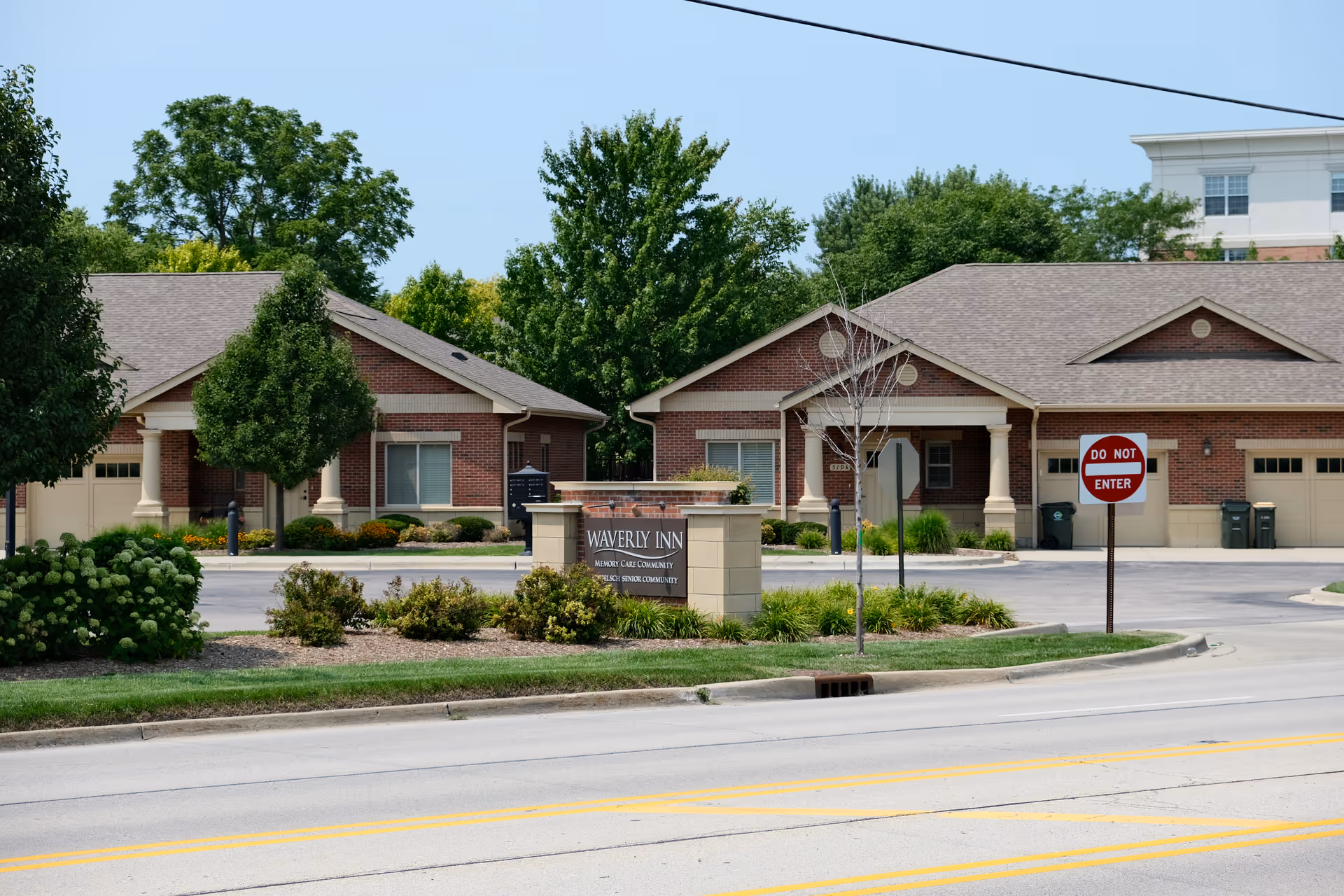Exterior view of Waverly Inn Memory Care Community showing brick buildings with garages, a landscaped area with bushes and trees, a sign with the facility name, and a Do Not Enter traffic sign near the driveway entrance.