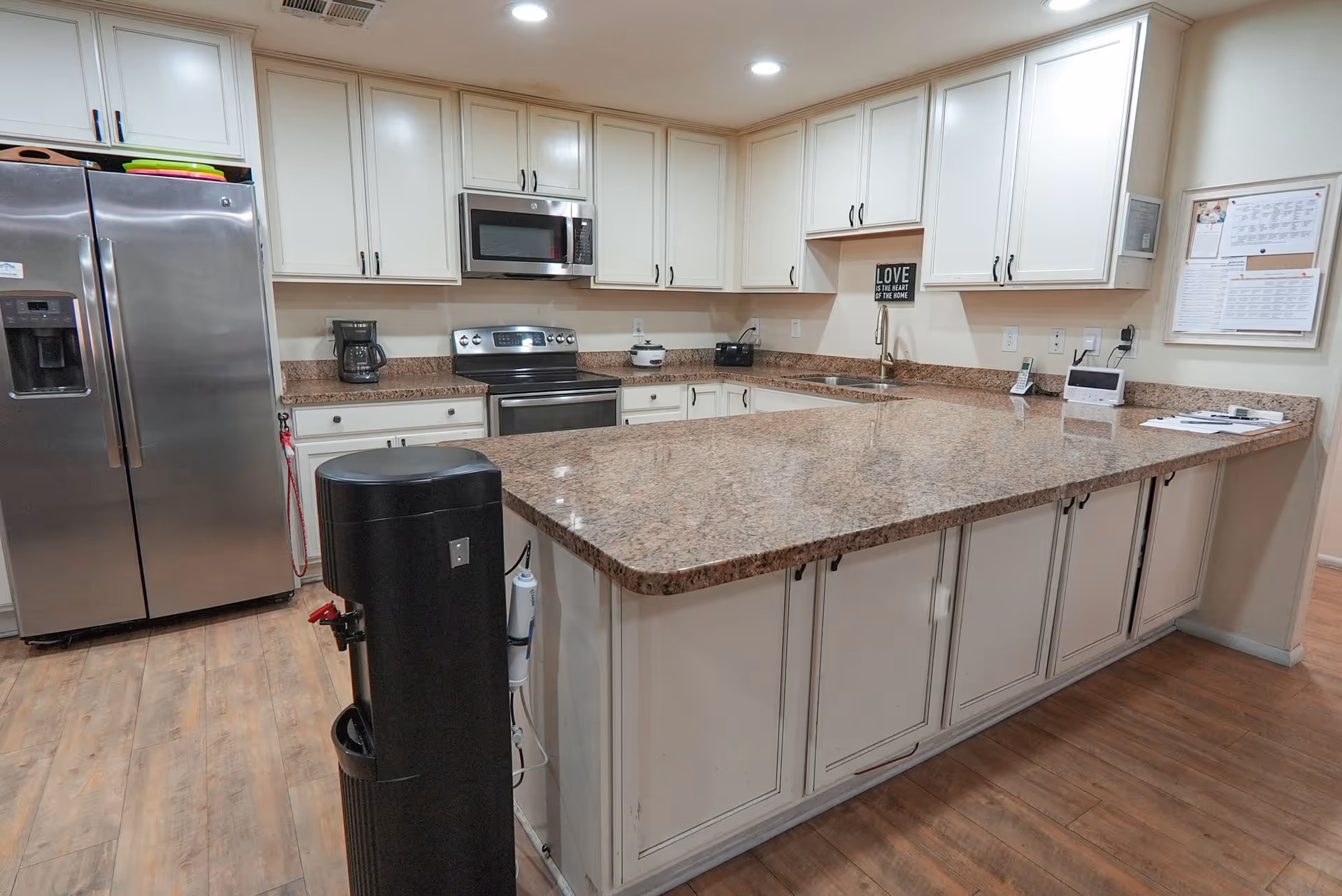 A spacious kitchen with white cabinets, granite countertops, and stainless steel appliances including a refrigerator, microwave, and stove. There is a large kitchen island in the center with additional storage underneath. The floor is wooden, and there is a black water dispenser in the foreground. A bulletin board and a small sign that reads 'LOVE is the heart of the home' are visible on the wall.