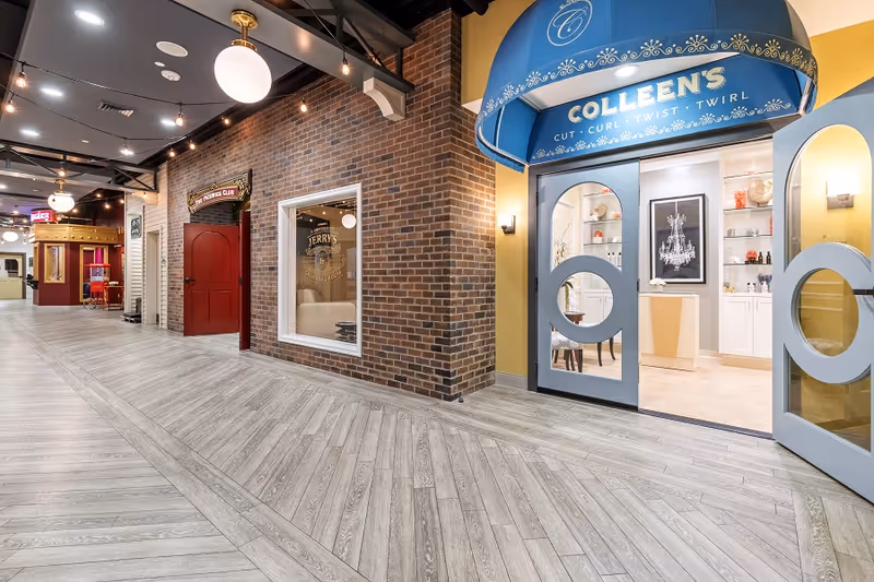 Interior hallway of a senior living facility featuring a salon named Colleen's with a blue awning and glass double doors, a brick wall with a window displaying Jerry's barbershop, and a popcorn club entrance with red doors further down the hall. The floor is light wood with a herringbone pattern and the ceiling has exposed beams and modern lighting fixtures.
