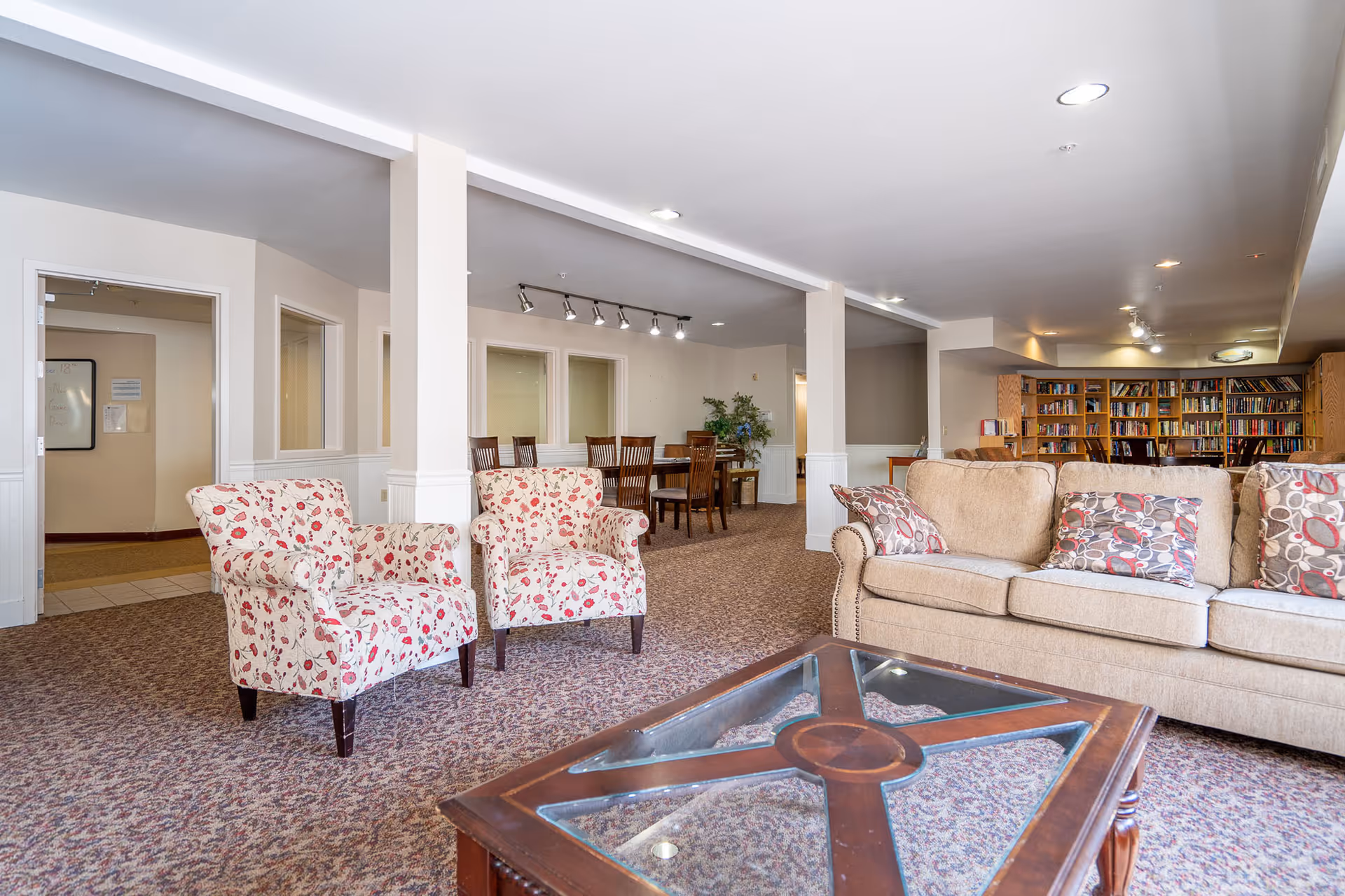 A spacious common area in a retirement facility featuring a beige sofa with patterned cushions, two floral armchairs, a glass-top wooden coffee table, a dining table with chairs in the background, and a large bookshelf filled with books along the back wall.