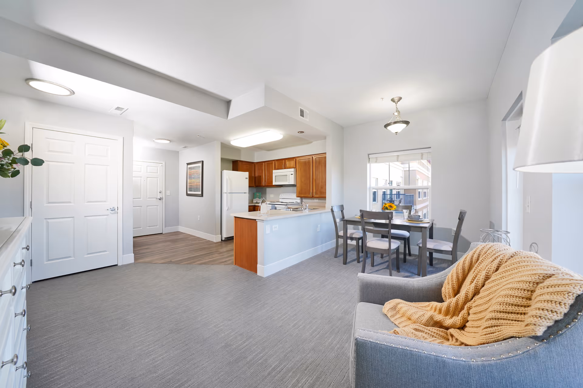 Bright and modern open-concept living space featuring a small kitchen with wooden cabinets, white appliances, and a breakfast bar. Adjacent to the kitchen is a dining area with a table set for four, and a cozy sitting area with a gray armchair draped with a beige knitted blanket. The room has light gray walls, carpeted flooring in the living and dining areas, and wood flooring near the entrance. A window lets in natural light, and the space is well-lit with ceiling lights.