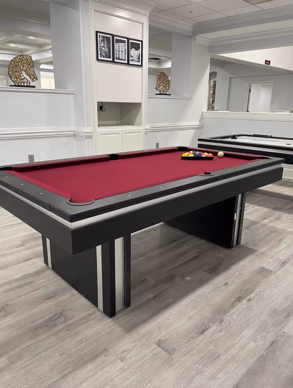 Interior room with a burgundy pool table set up with billiard balls and a cue ball, next to an air hockey table. The room has light gray wood flooring, white walls, and decorative horse head sculptures on a ledge.