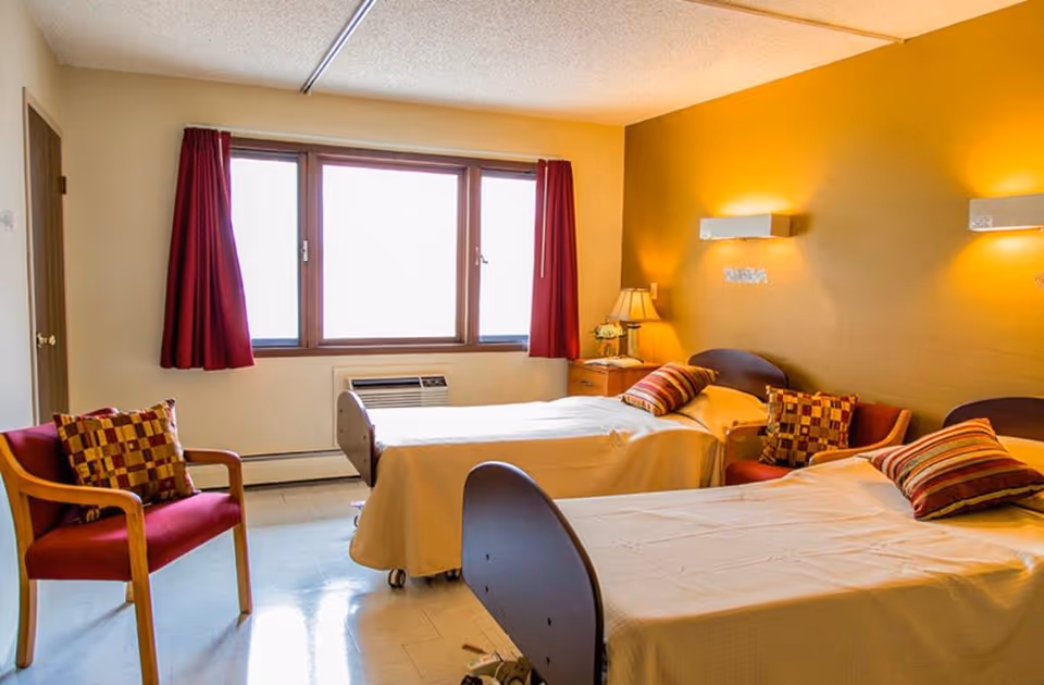 A bright room with two single beds covered in beige bedspreads, each with striped and checkered pillows. There is a wooden chair with a red cushion and a checkered pillow near the window, which has red curtains. A nightstand with a lamp and a small flower arrangement is placed between the beds. The walls are painted yellow and beige, and the room has a clean, simple, and cozy atmosphere.
