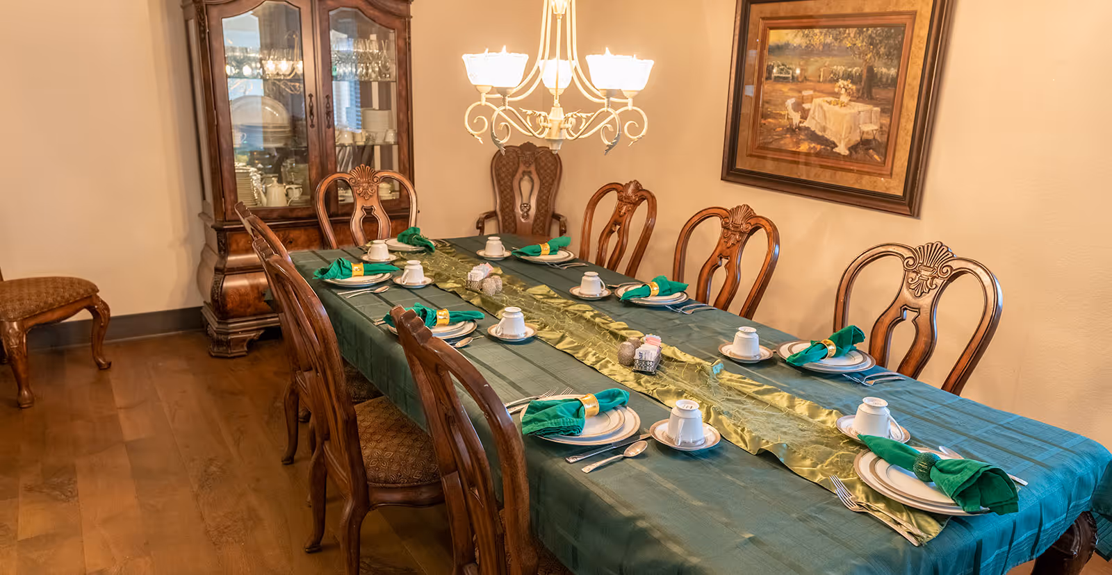 A formal dining room with a long wooden table covered with a green tablecloth and a gold table runner. The table is set with white plates, cups, silverware, and green cloth napkins with gold napkin rings. There are eight wooden chairs around the table. A wooden china cabinet with glass doors displaying dishes is against the wall. A chandelier with multiple lights hangs above the table. A framed painting of an outdoor dining scene is on the wall.