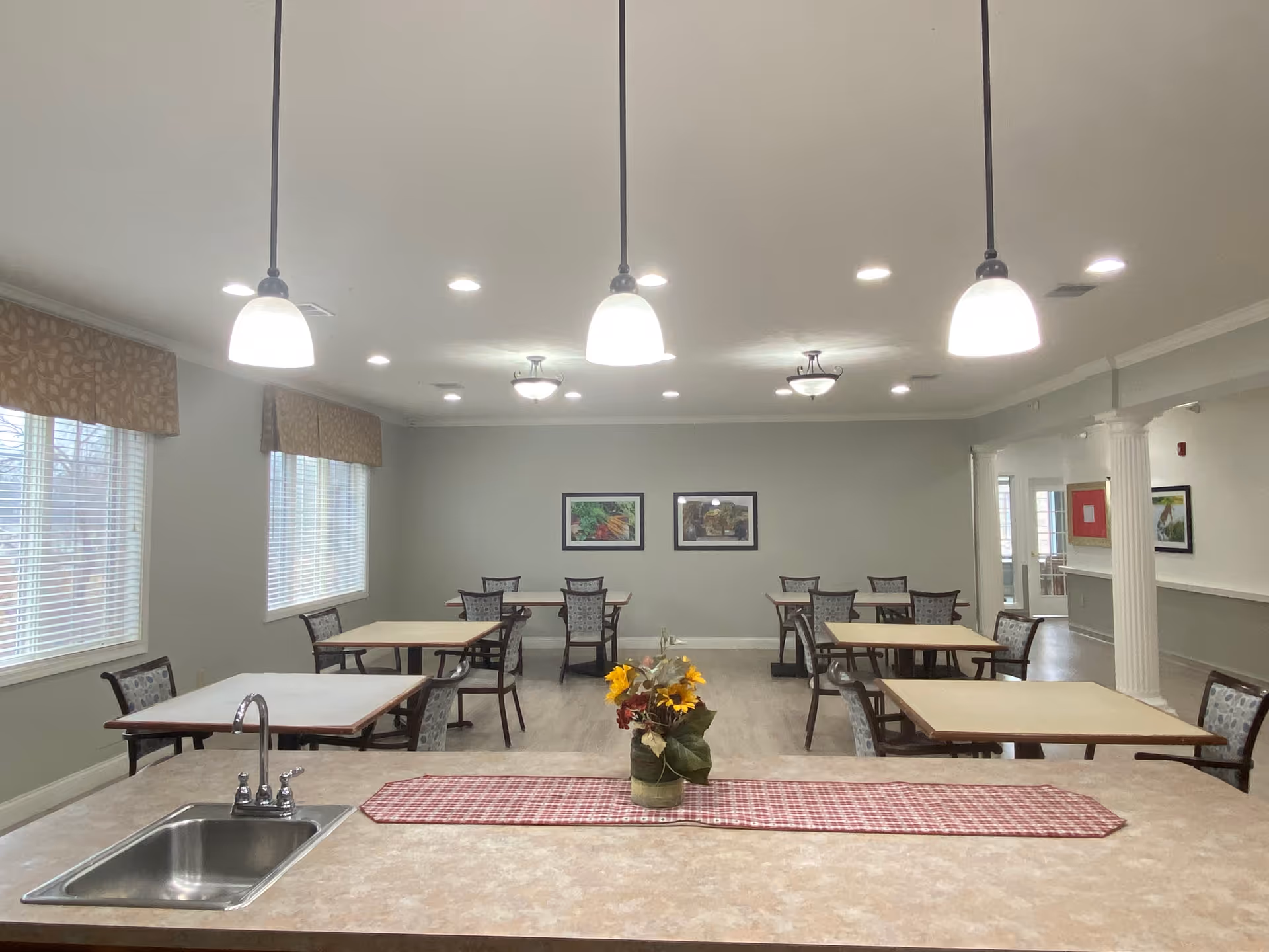 A dining room with several square tables and chairs arranged neatly. The room has large windows with patterned valances, overhead pendant lights, and framed pictures on the far wall. In the foreground, there is a countertop with a sink and a small vase of flowers on a red checkered runner.