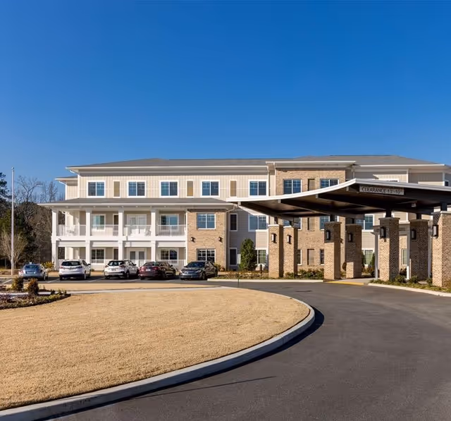 Front exterior of a three-story senior living building with a covered porte-cochere, parked cars, and a curved driveway under a clear blue sky.