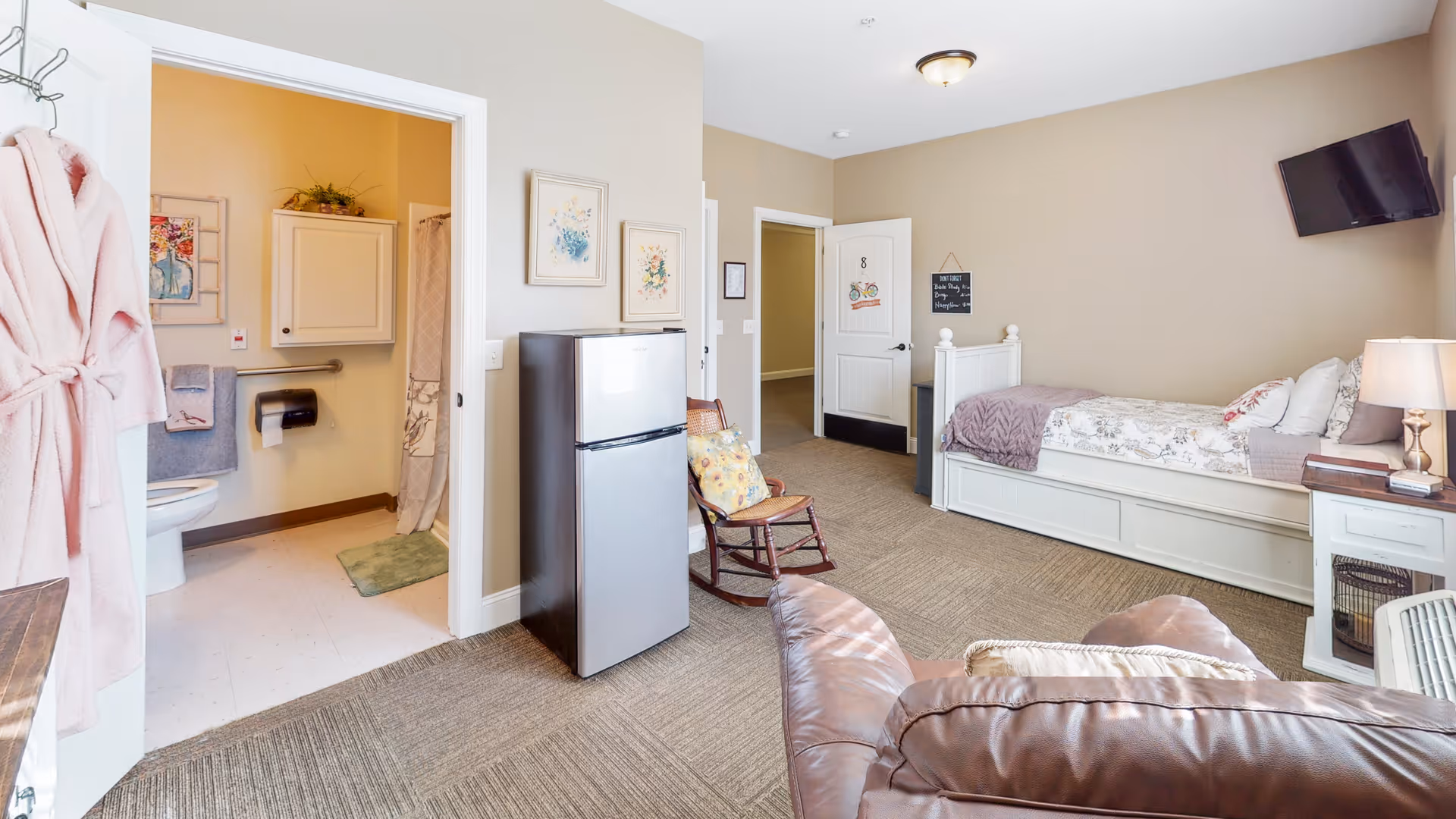 Furnished senior living studio with a bed, reclining chair, small refrigerator, wall-mounted TV and an open bathroom.