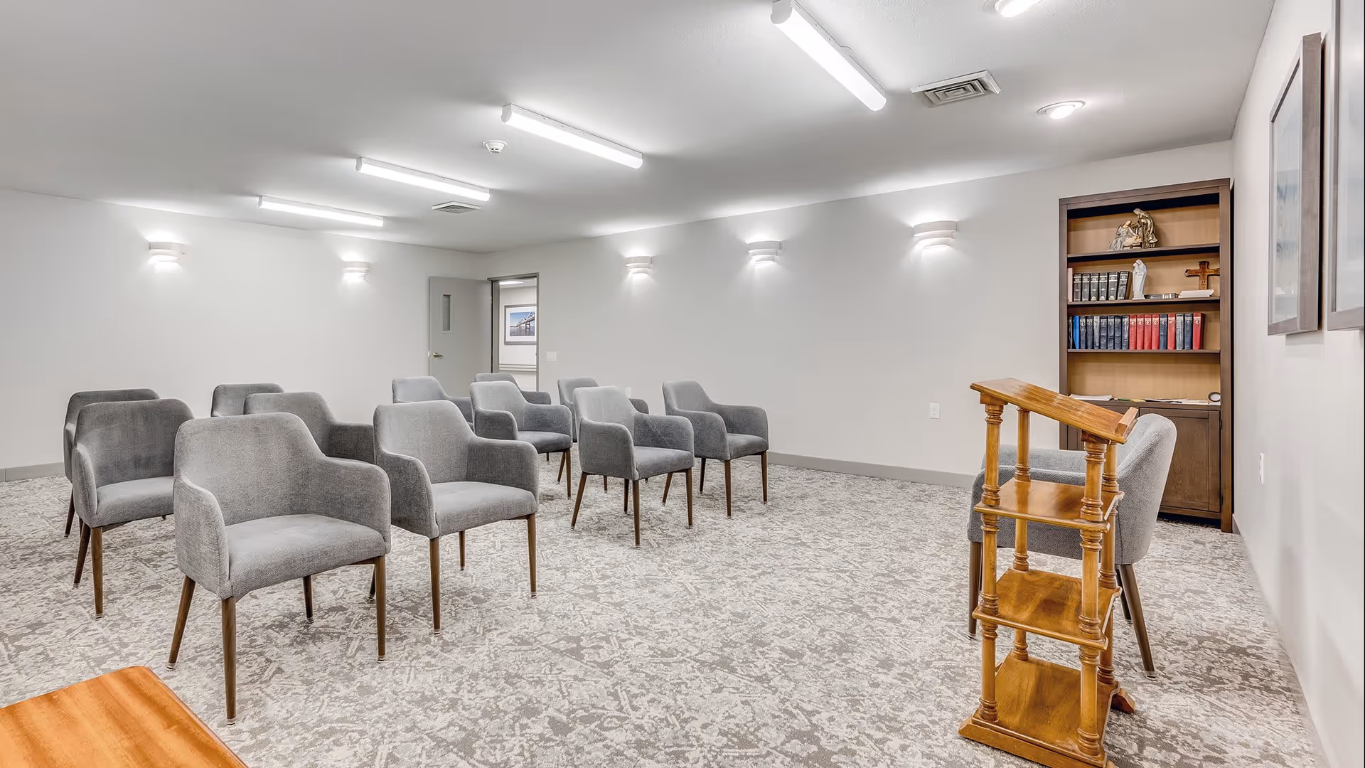 A small meeting or presentation room with several gray upholstered chairs arranged in rows facing a wooden podium. The room has light gray walls, a patterned carpet, wall-mounted lights, and a bookshelf with books and decorative items in the corner.