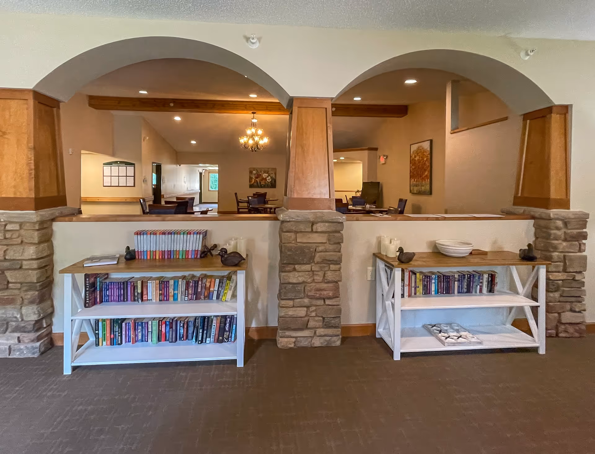 Interior view of a senior living facility showing two white bookshelves filled with books and decorative items, positioned against a half wall with stone pillars and wooden arches. In the background, there are tables and chairs under a chandelier, with paintings on the walls.