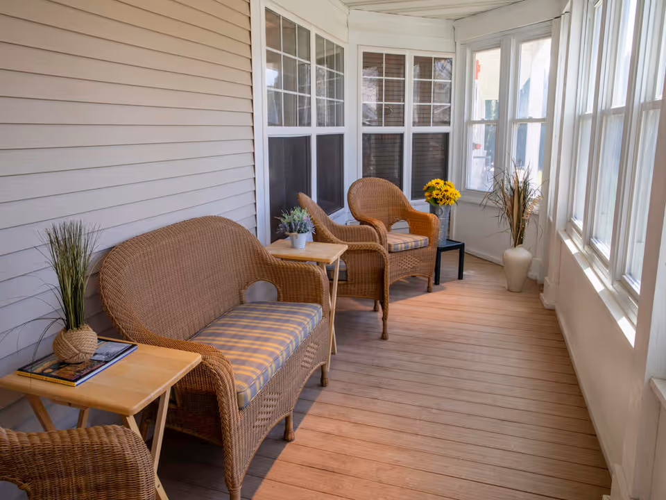 A bright enclosed porch area with wooden flooring and beige siding walls, furnished with wicker chairs and a loveseat with plaid cushions. Small wooden side tables hold potted plants and books, and there are large windows letting in natural light. A vase with yellow flowers and a tall white vase with decorative grass are placed near the windows.