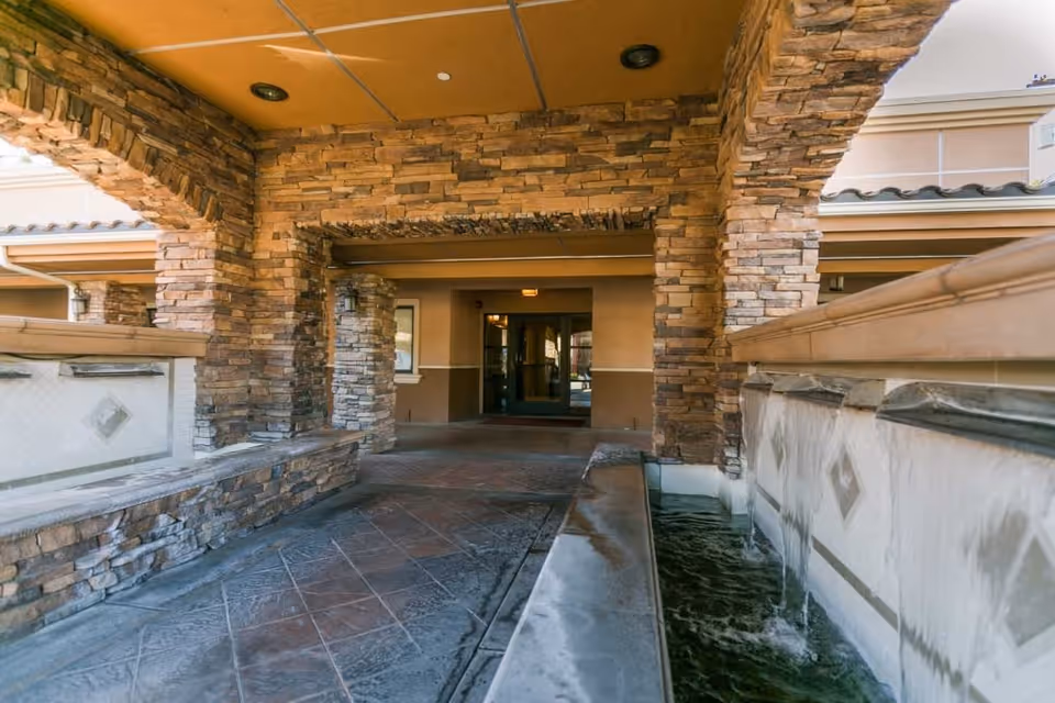 Covered outdoor walkway with stone pillars and walls, featuring a water fountain with flowing water on the right side, leading to a glass door entrance of a building.