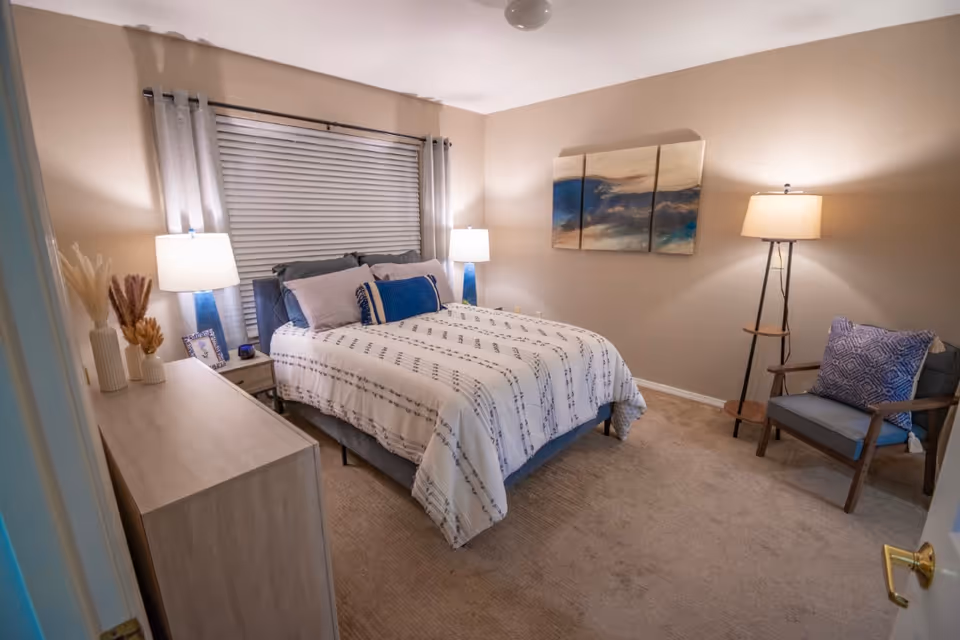 A cozy bedroom with a neatly made bed featuring white and blue patterned bedding. There are two bedside tables with matching lamps on either side of the bed. A window with closed blinds and light gray curtains is behind the bed. A wooden dresser with decorative vases and dried plants is on the left side of the room. On the right side, there is a wooden armchair with a blue cushion and a floor lamp beside it. A three-panel abstract painting hangs on the wall above the chair. The room has beige walls and carpeted flooring.