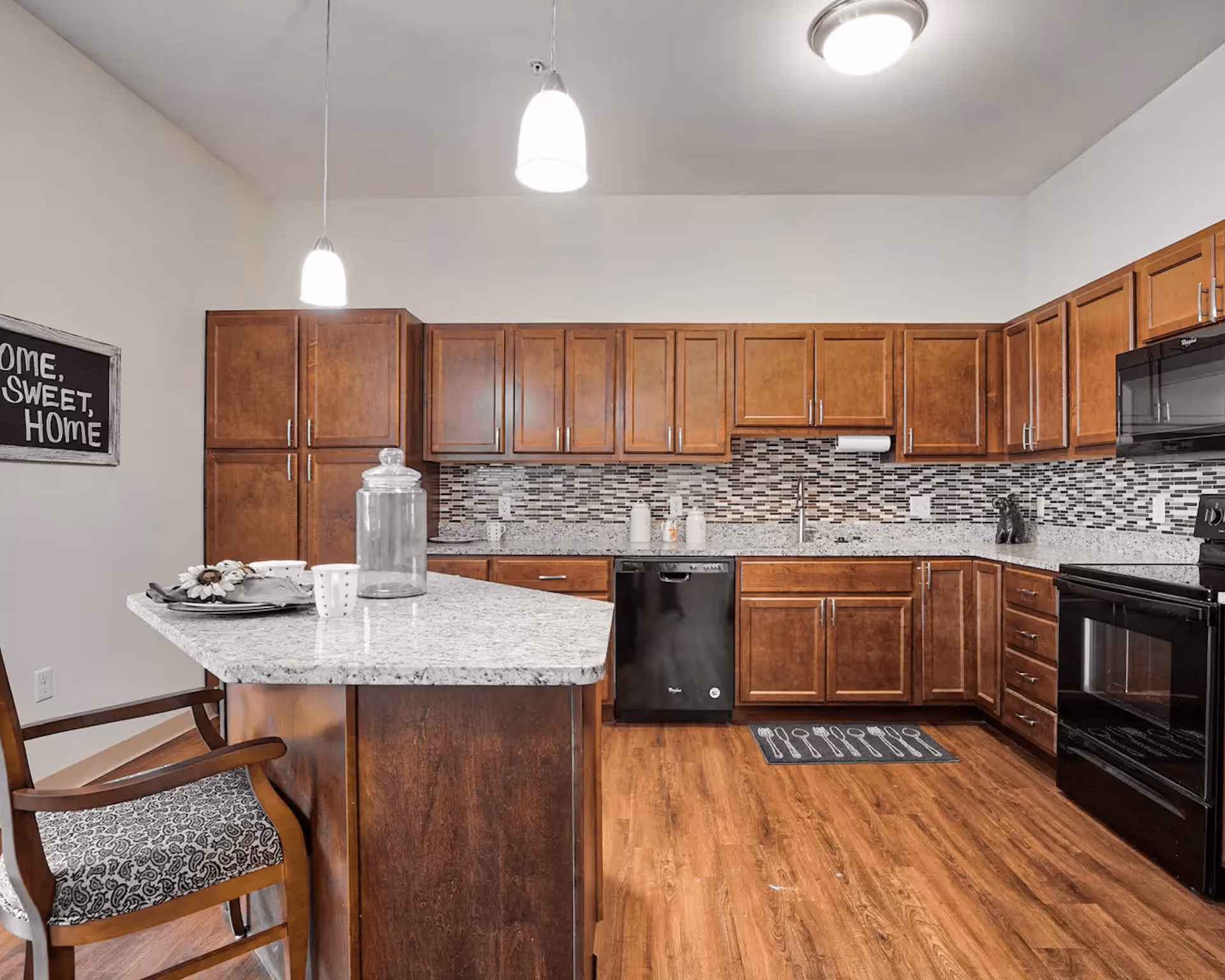 A modern kitchen with wooden cabinets, granite countertops, and a tiled backsplash. The kitchen features a black dishwasher, black oven, and a kitchen island with a chair. Two pendant lights hang from the ceiling, and a decorative sign on the wall reads 'Home, Sweet, Home.'