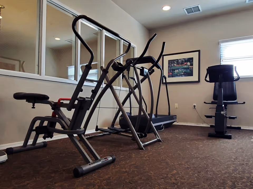 A small fitness room with exercise equipment including a recumbent bike, elliptical machines, and a treadmill. The room has carpeted flooring, a window with blinds, a framed picture on the wall, and a mirrored wall reflecting the equipment.