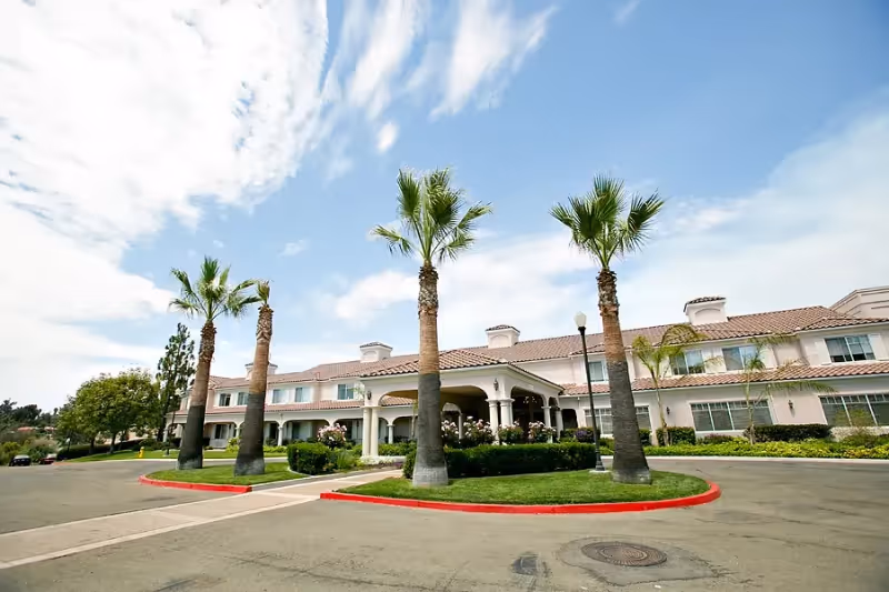 Exterior view of a two-story senior living facility with a tiled roof, surrounded by palm trees and landscaped greenery under a partly cloudy sky.