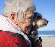 An elderly woman with short gray hair wearing a red sweater and a light pink scarf is holding a small dog while looking out towards the ocean on a beach.