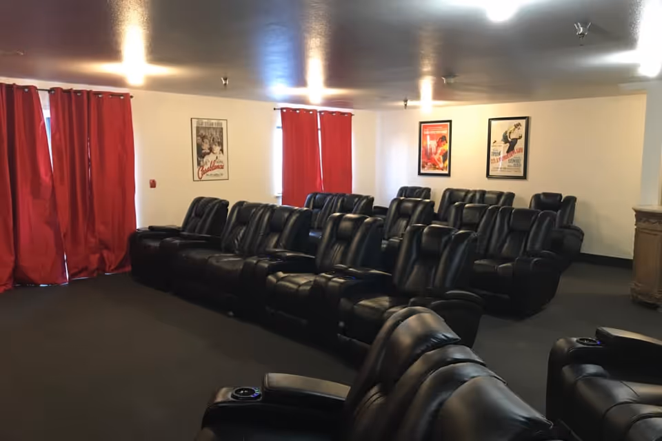 A small theater room with three rows of black leather recliner chairs facing a screen or stage area. The walls are white with three framed vintage movie posters and red curtains covering the windows.