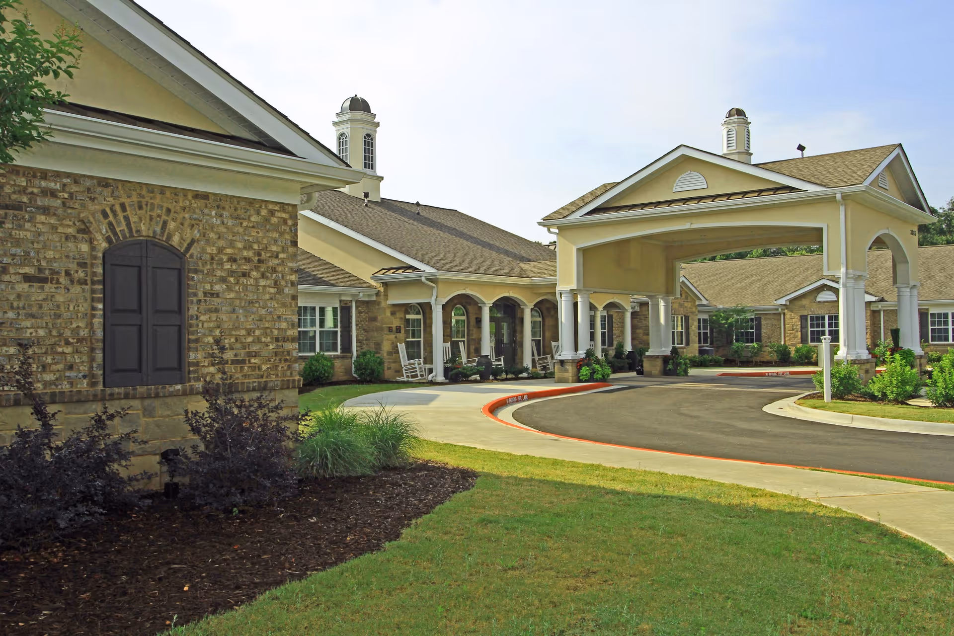 Front exterior of a senior living facility showing a covered entrance (porte-cochere), brick facade, and circular driveway.