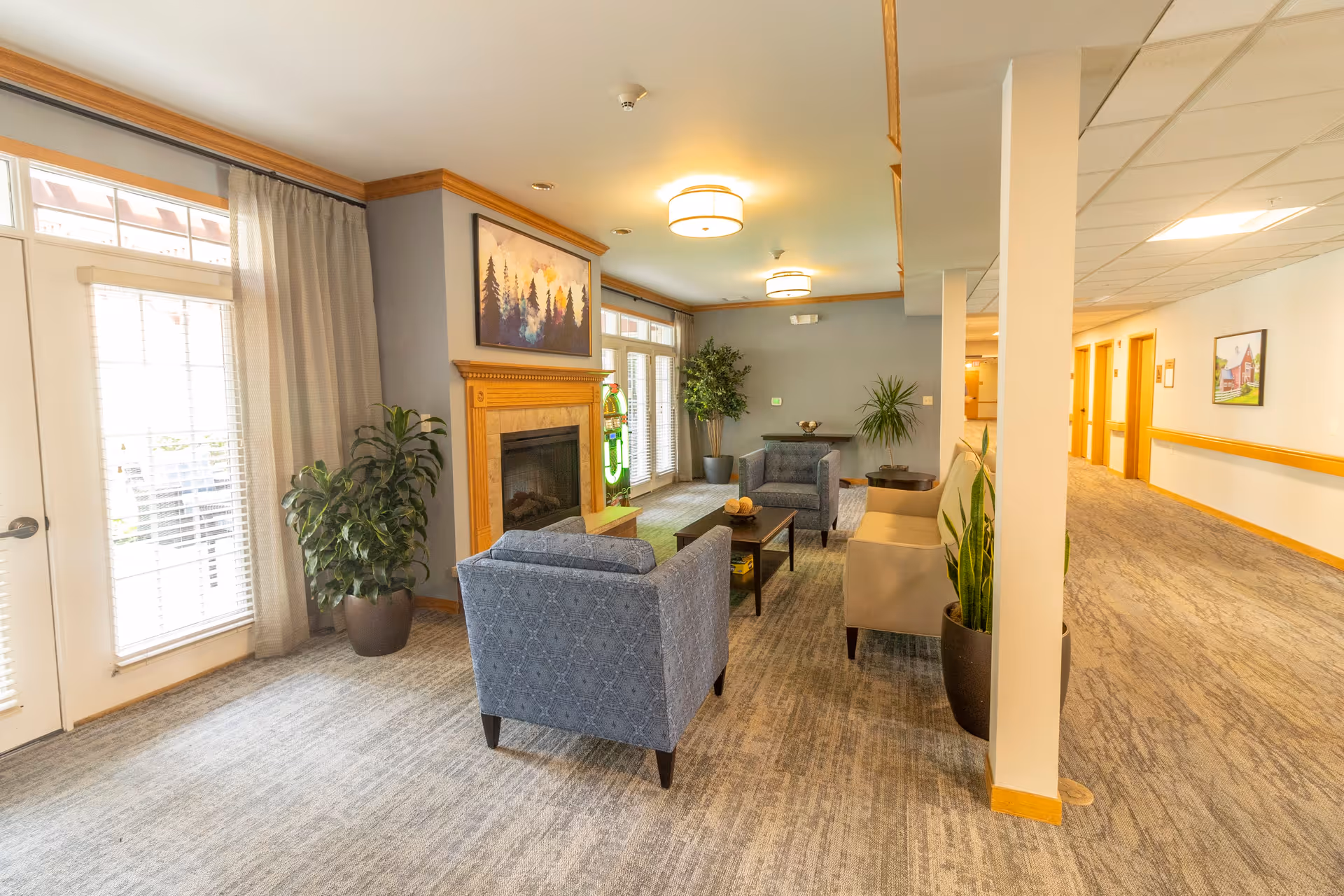 A cozy living room area in a senior living facility with two blue armchairs, a beige sofa, a wooden coffee table, and a fireplace with a painting of trees above it. There are large windows with sheer curtains letting in natural light, several potted plants, and a hallway leading to other rooms.