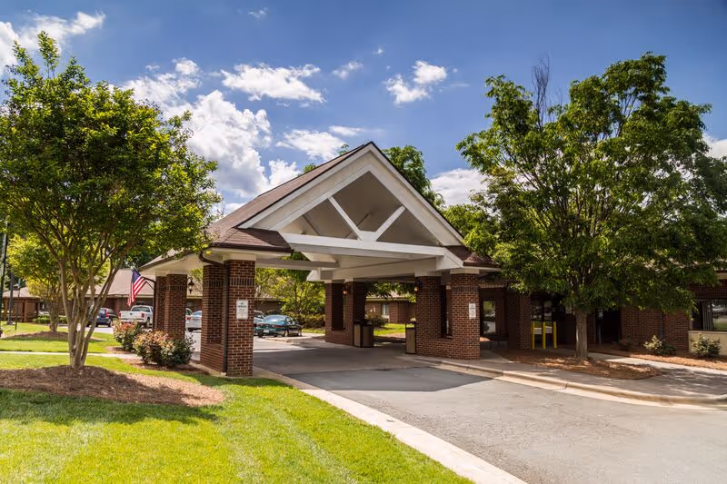 Covered brick entrance (porte-cochère) of a senior care facility with a driveway, landscaped lawn, and trees under a partly cloudy sky.