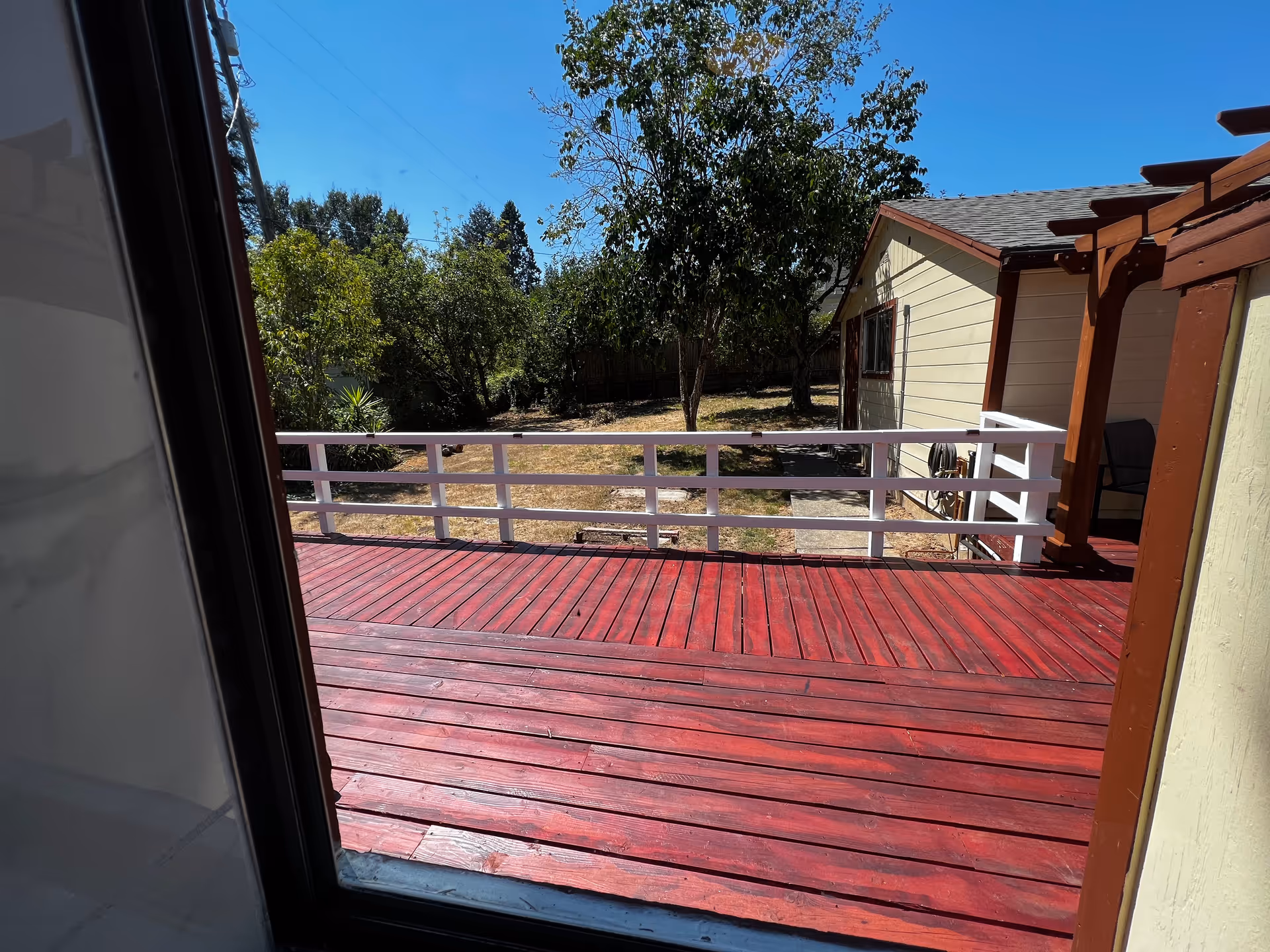 View through a window showing a red wooden deck with white railing, a small yard with trees and grass, and a light yellow building with brown trim under a clear blue sky.