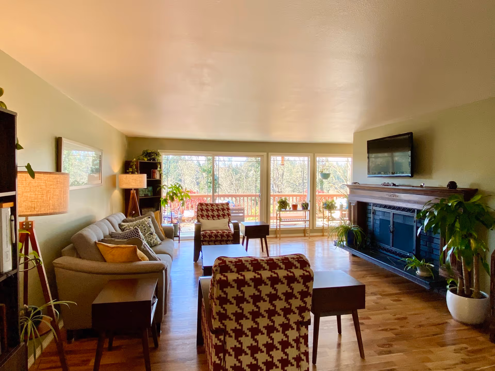 A bright and cozy living room with wooden flooring, a beige sofa with multiple cushions, two red and white patterned armchairs, a coffee table, side tables, and several potted plants. There is a fireplace with a mounted flat-screen TV above it on the right wall. Large windows and a sliding glass door at the far end let in natural light and offer a view of an outdoor deck and trees.