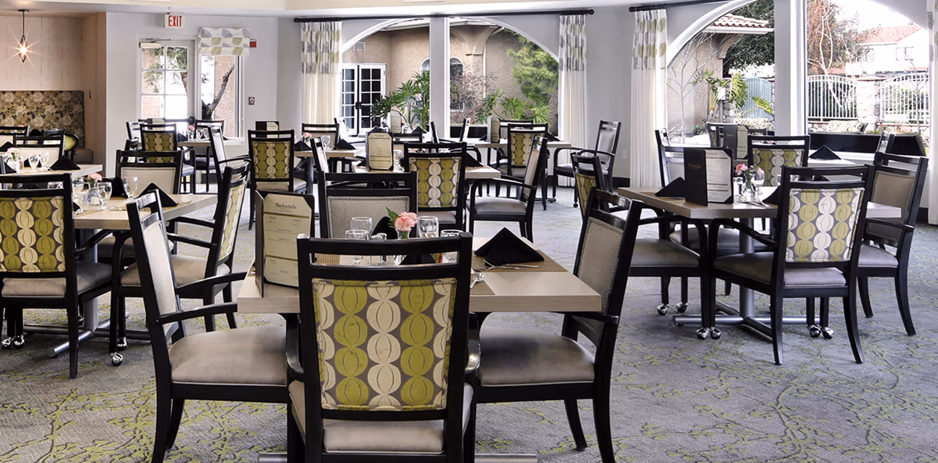 A bright and spacious dining room with multiple tables and chairs arranged neatly. The chairs have patterned backs with green and beige colors. Each table is set with glasses, napkins, and menus. Large windows with white curtains allow natural light to fill the room, and there is a view of an outdoor area with greenery.