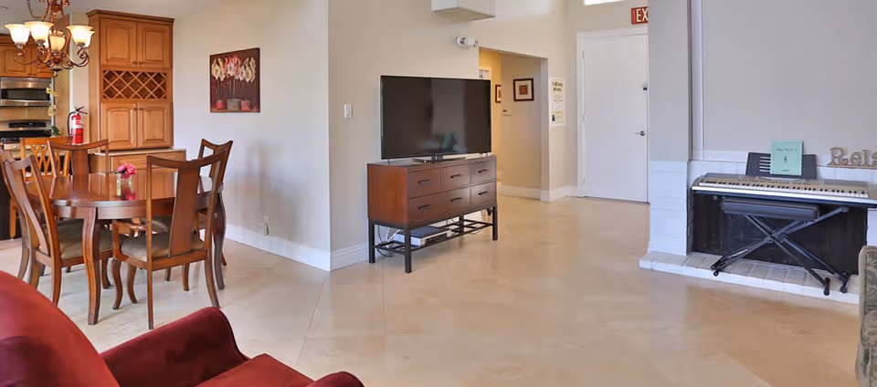 Bright common living area with a dining table and chairs, a TV on a stand, and an electronic keyboard by a fireplace.