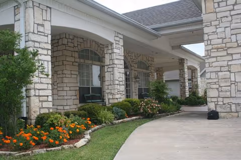 Exterior view of a stone building with large pillars and arched windows, a covered driveway, and landscaped garden beds with green shrubs and orange flowers.