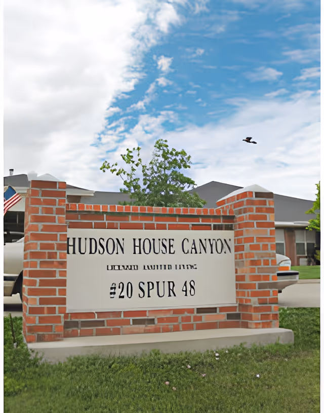 A brick sign for Hudson House Canyon, a licensed living facility, located on a grassy area with a partly cloudy blue sky in the background. A vehicle and part of a building are visible behind the sign.