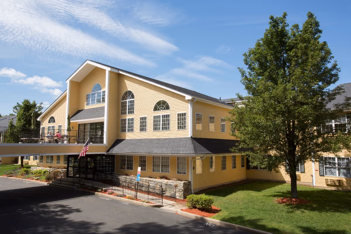 Exterior view of a yellow senior living facility building with multiple large windows, a ramp and stairs leading to the entrance, an American flag near the entrance, and a tree on the right side under a partly cloudy blue sky.