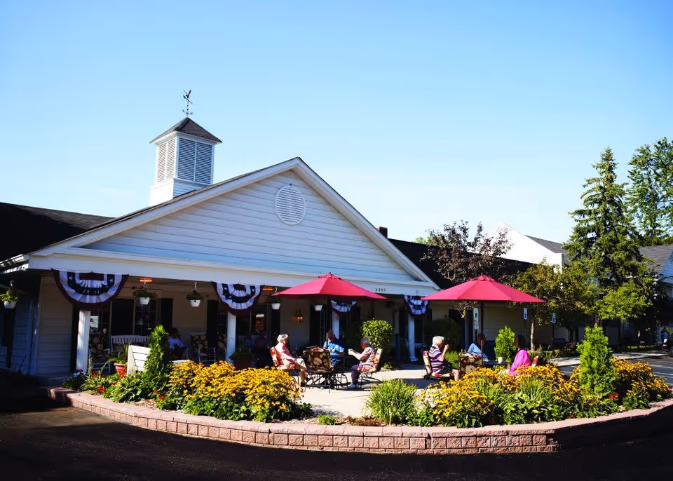 Outdoor patio area at American House Elmwood with several elderly people sitting and chatting under red umbrellas. The building has white siding with patriotic bunting decorations and a cupola on the roof. There are flower beds with yellow flowers and greenery surrounding the patio.