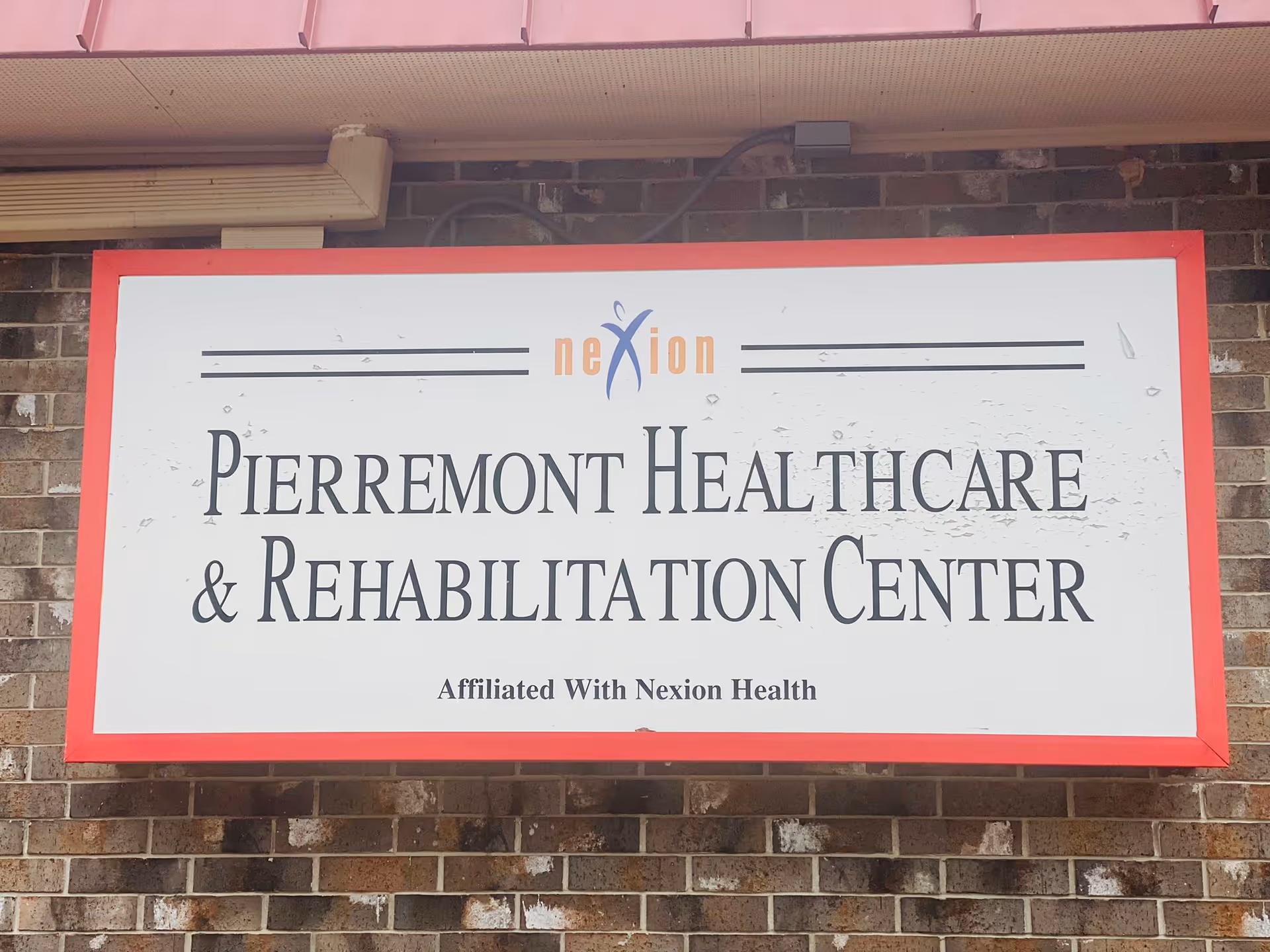 A rectangular sign mounted on a brick wall with a red border. The sign reads 'Pierremont Healthcare & Rehabilitation Center' with a smaller text below stating 'Affiliated With Nexion Health'. The word 'nexion' is stylized with a blue and orange logo above the main text.