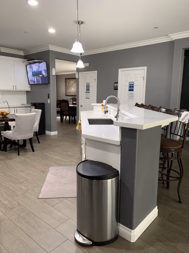 Interior view of a modern kitchen area in an elderly care facility. The image shows a white countertop with a sink and a stainless steel trash can in front. There are several bar stools lined up along the counter. In the background, there is a dining area with a round table and upholstered chairs, a wall-mounted TV, and two closed doors with restroom signs. The walls are painted gray and the floor has wood-like tiles.