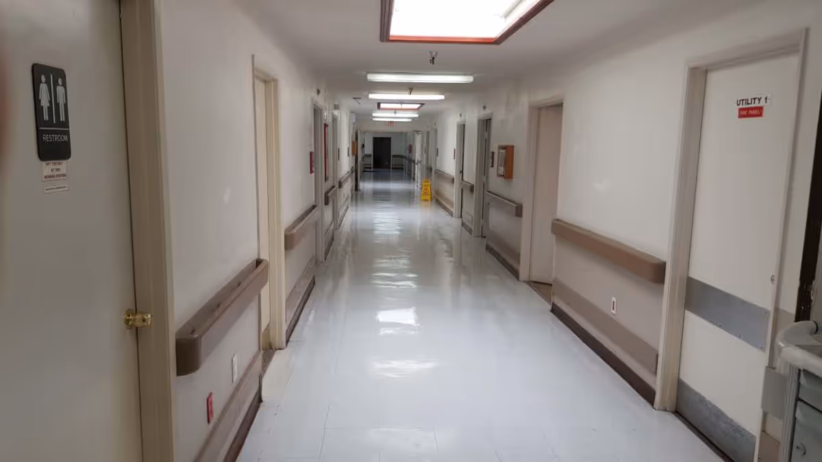 A long, clean hospital hallway with white walls and floor, handrails on both sides, multiple closed doors, and ceiling lights. A restroom sign is visible on the left door, and a utility room door is on the right.