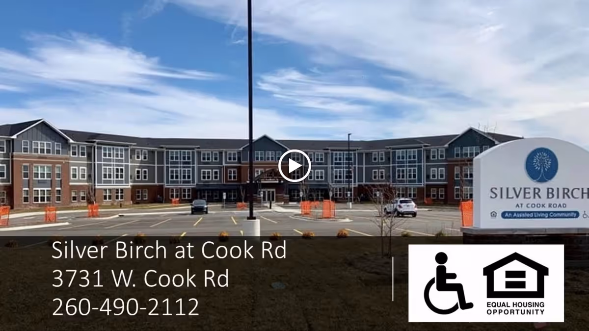 Exterior view of Silver Birch at Cook Road, a large three-story assisted living community building with a parking lot in front and a sign displaying the facility name and contact information.