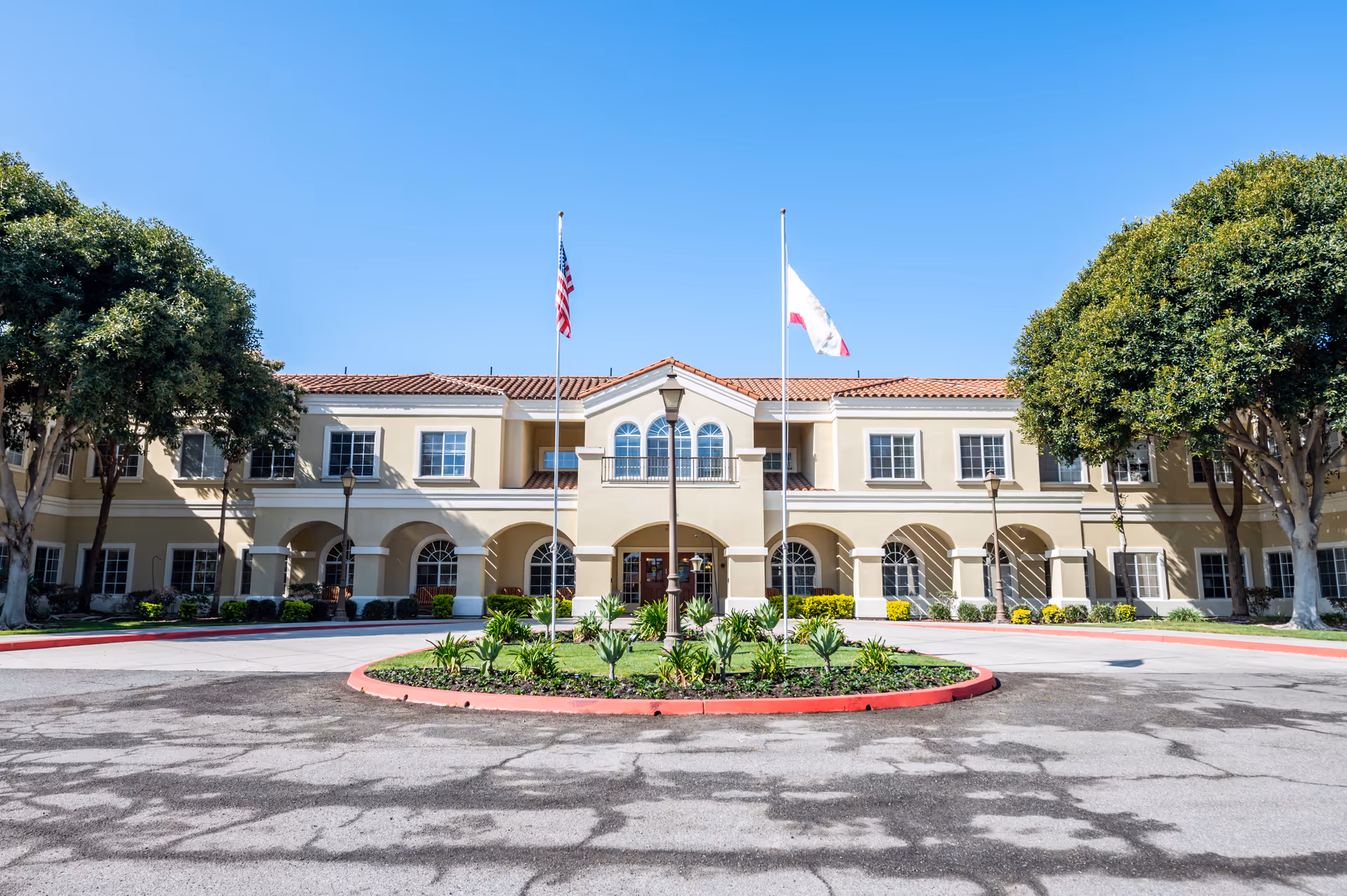 Front exterior view of Capistrano Senior Living building with beige walls, red tile roof, two flagpoles with American and California flags, a circular driveway with landscaped greenery in the center, and large trees on either side under a clear blue sky.