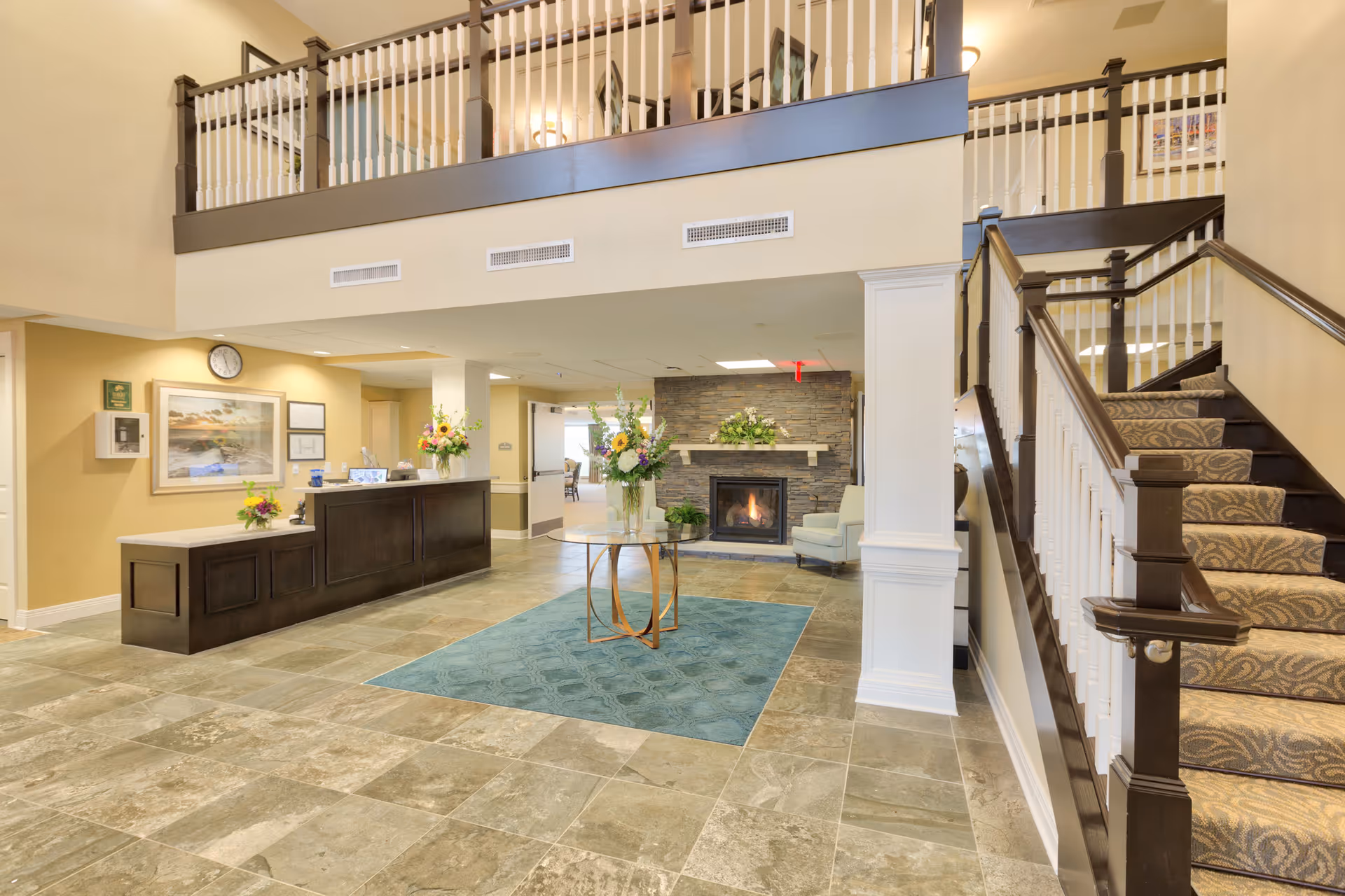 A spacious and well-lit senior living facility lobby with a dark wood reception desk on the left, a stone fireplace with a floral arrangement on the mantel in the center, and a glass table with a flower vase on a blue rug in front of the fireplace. To the right is a carpeted staircase with dark wood handrails and white balusters leading to an upper floor with a balcony overlooking the lobby.