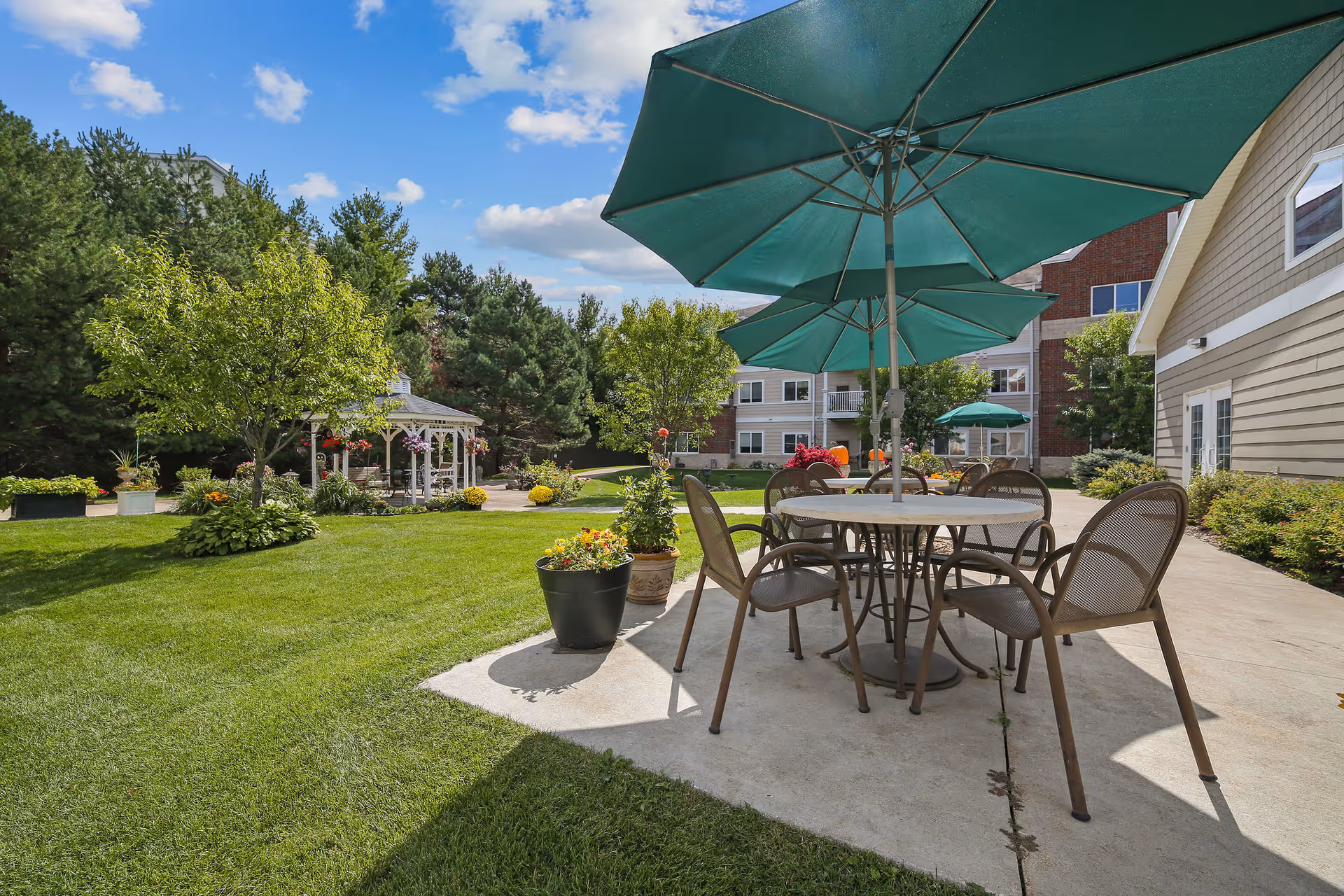 Patio with a round table, chairs and large green umbrellas on a concrete slab overlooking a grassy courtyard, gazebo and surrounding buildings.