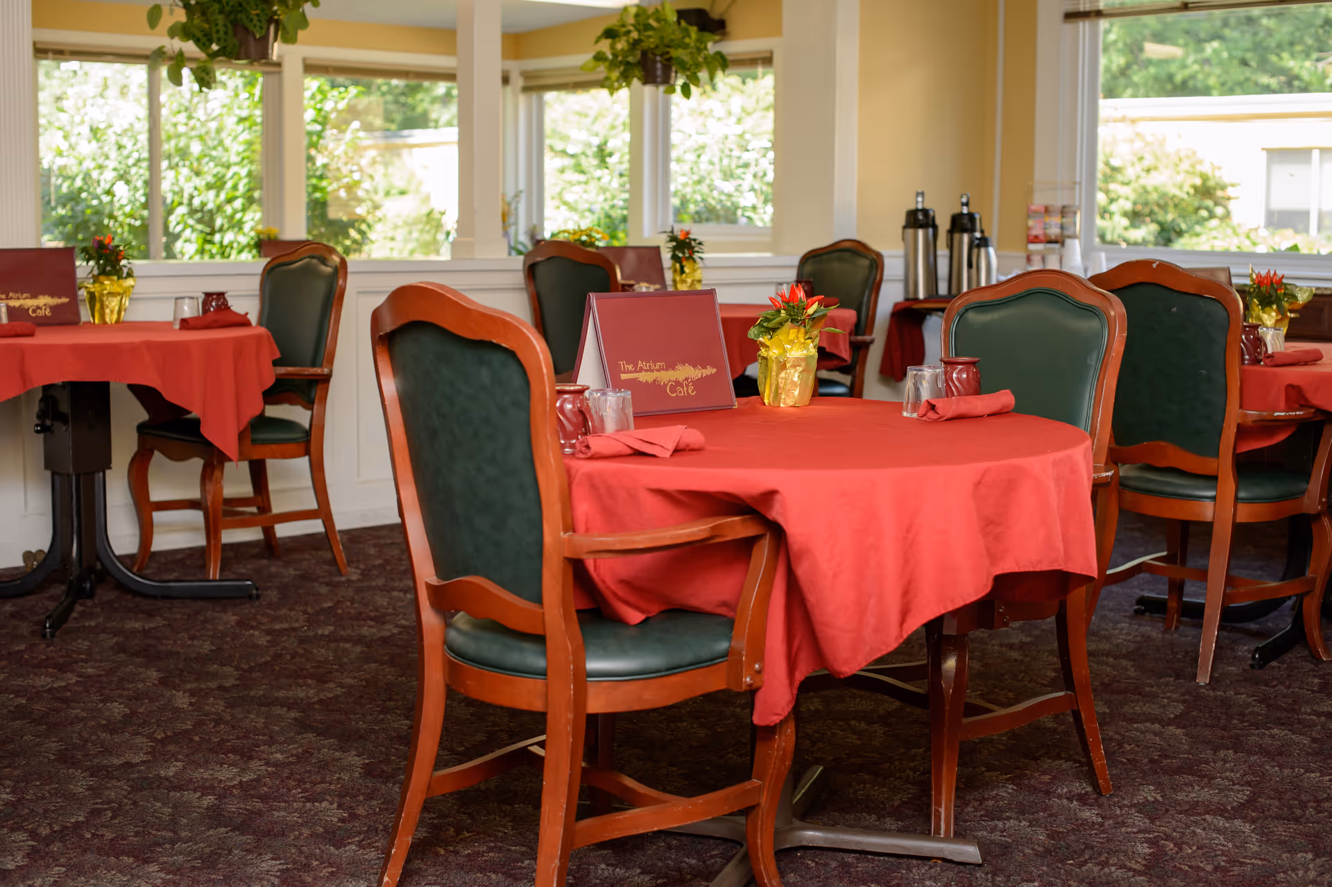 Dining area with round tables covered in red tablecloths, green cushioned wooden chairs, small flower arrangements in gold wrapping, and menus on the tables. Large windows allow natural light and show greenery outside.