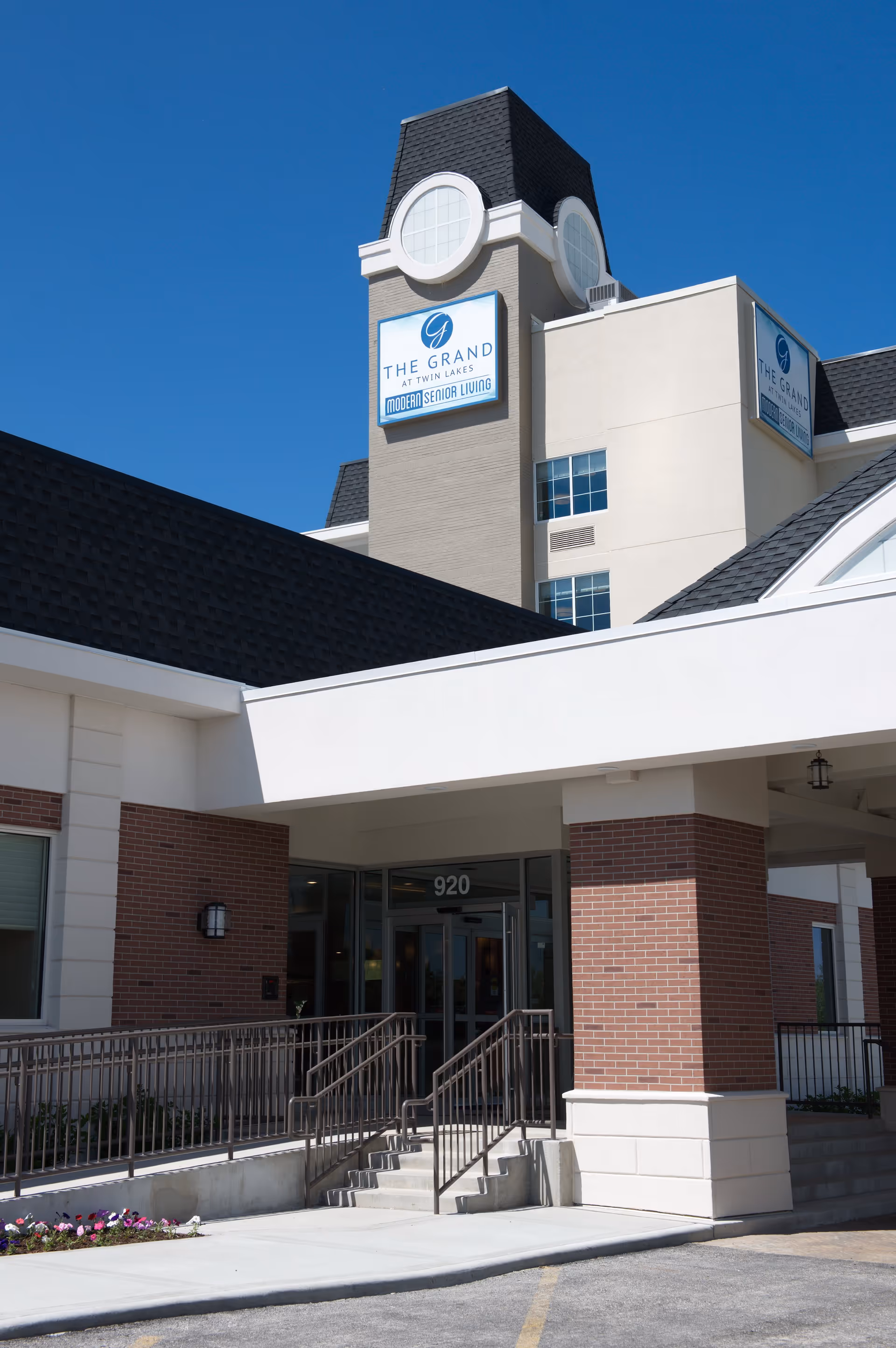 Entrance of The Grand at Twin Lakes senior living facility showing a brick and beige building with a covered entryway, stairs, and a ramp. The building has a tower with circular windows and signs displaying the facility name and 'Modern Senior Living' under a clear blue sky.