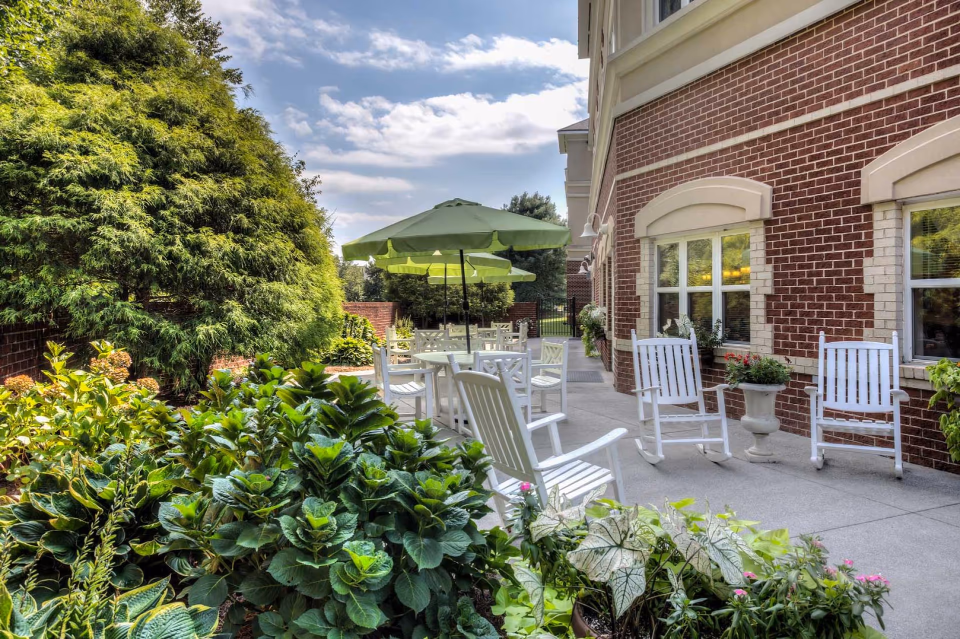 Outdoor patio area at PruittPlace - Winston Salem with white rocking chairs and tables under green umbrellas, surrounded by lush greenery and plants next to a red brick building with windows.