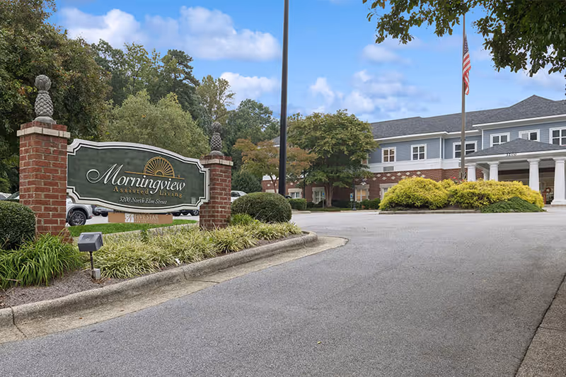 Entrance to Morningview Assisted Living facility with a large sign on brick pillars, a driveway leading to the building, landscaped bushes and trees, and an American flag on a flagpole in front of the building under a partly cloudy sky.