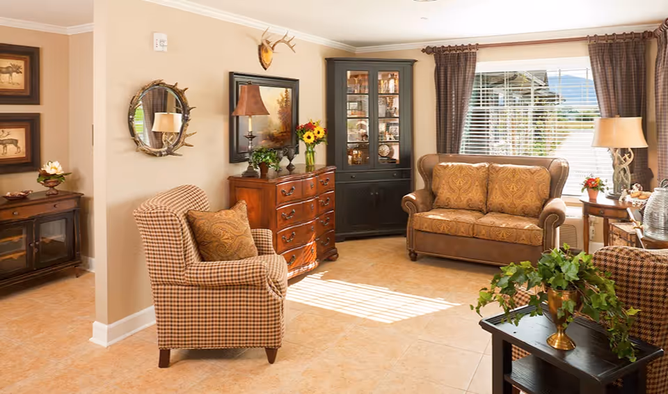 A cozy living room with a patterned armchair and matching loveseat, a wooden chest of drawers with a table lamp and a vase of flowers, a black display cabinet with glass doors, a round mirror with a rustic frame, framed artwork on the walls, and a window with brown curtains letting in natural light. There are also small tables with decorative plants and lamps.
