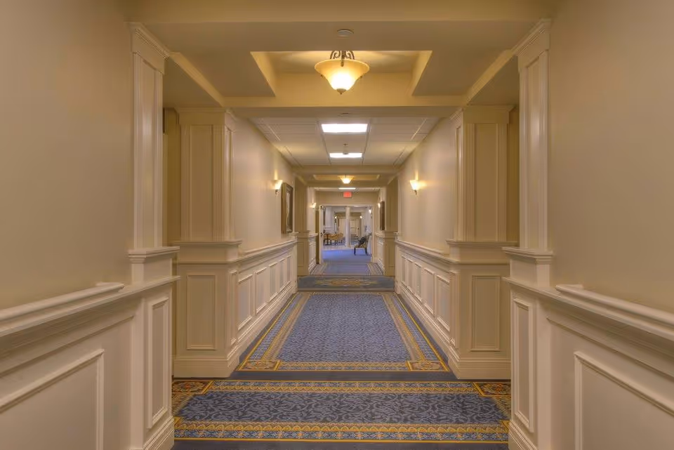 Long, well-lit interior hallway with blue patterned carpet, white wainscoting, and ceiling lights.