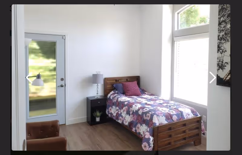 A small bedroom with a single wooden bed covered with a floral patterned bedspread and a purple pillow. Next to the bed is a black nightstand with a gray lamp and a small potted plant. There is a large window letting in natural light and a glass door with a white frame. A brown upholstered chair is partially visible in the foreground.