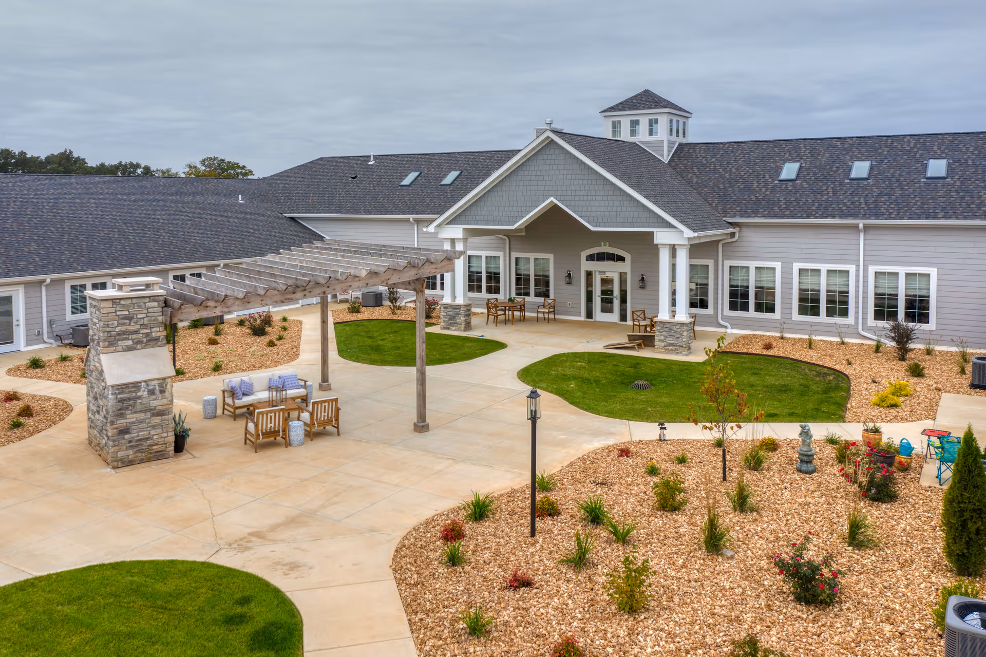 Front entrance and landscaped courtyard of a senior living facility with a pergola, outdoor seating, and decorative plant beds.