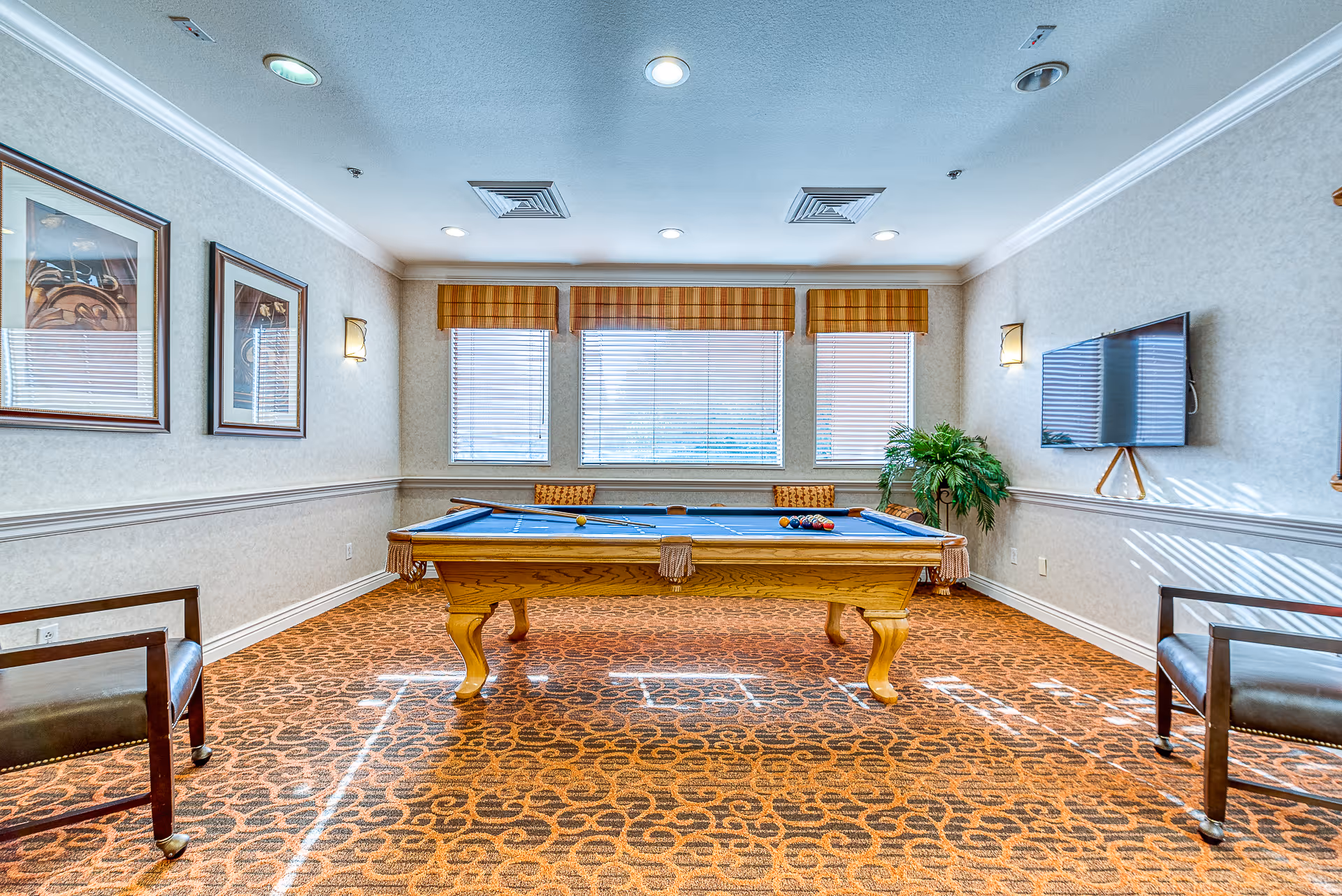 A well-lit game room with a blue felt pool table in the center, two wooden chairs with leather seats on either side, three windows with blinds and valances, framed artwork on the left wall, a flat-screen TV mounted on the right wall, and a potted plant in the corner.