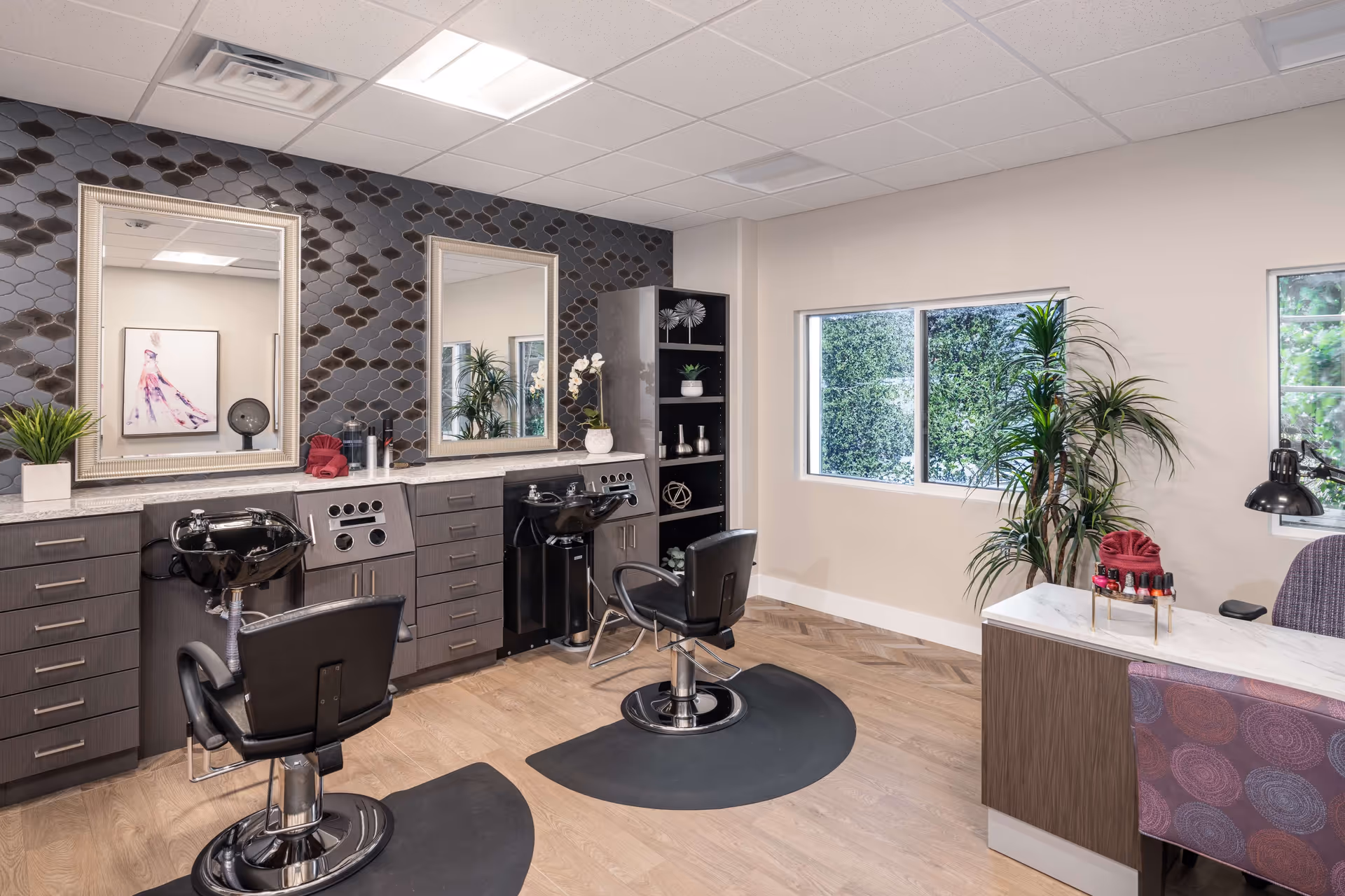 Bright salon-style interior with two styling chairs and wash sinks in front of mirrors, cabinets, and potted plants.