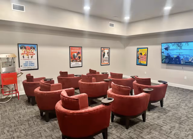 A small movie theater room with red upholstered chairs arranged in rows facing a wall-mounted flat screen TV. The walls are decorated with framed classic movie posters, and there is a red popcorn machine on a stand in the corner.